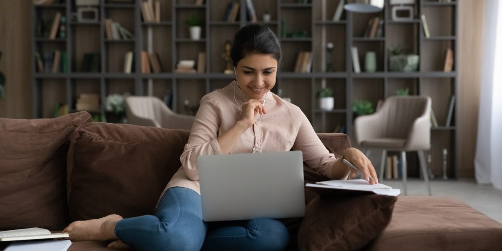 Smiling Indian woman in earphones studying online, using laptop, sitting on couch at home, motivated positive young female student watching webinar, involved in internet lesson, taking notes